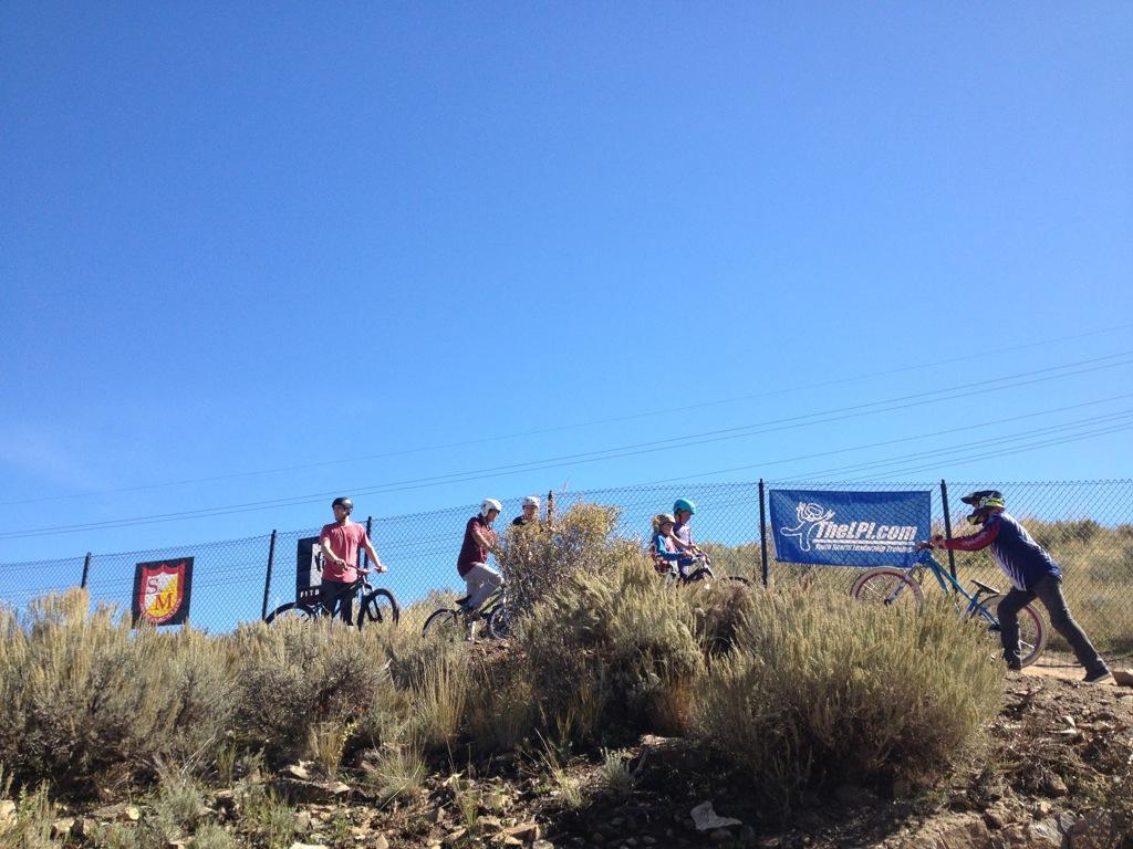 A group of five individuals, wearing helmets and casual clothing, are standing near their bicycles on a hillside with low shrubbery. The sky is clear and blue, and there is a fence in the background with two banners visible, one featuring a logo and the other displaying a website. The scene captures a moment related to outdoor biking activities. PC Dirt Jumps mountain bike trail.
