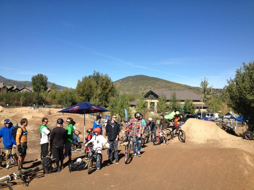 A gathering of mountain bikers in protective gear, standing on a dirt jump course surrounded by trees and residential buildings under a clear blue sky. Some riders are socializing while others prepare their bikes, with a banner and tents visible in the background. PC Dirt Jumps mountain bike trail.