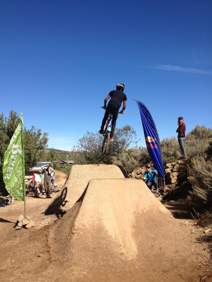 A mountain biker in a black shirt and helmet performs a jump over a dirt ramp at a biking event. Two flags are visible in the background, and spectators can be seen watching the action amidst a clear blue sky. PC Dirt Jumps mountain bike trail.