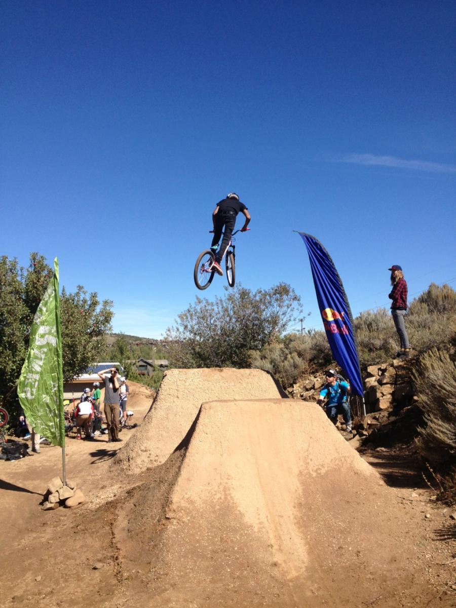 A cyclist performing a jump over dirt ramps at a biking event, with a clear blue sky in the background. Spectators can be seen watching the action, and banners are positioned nearby. PC Dirt Jumps mountain bike trail.