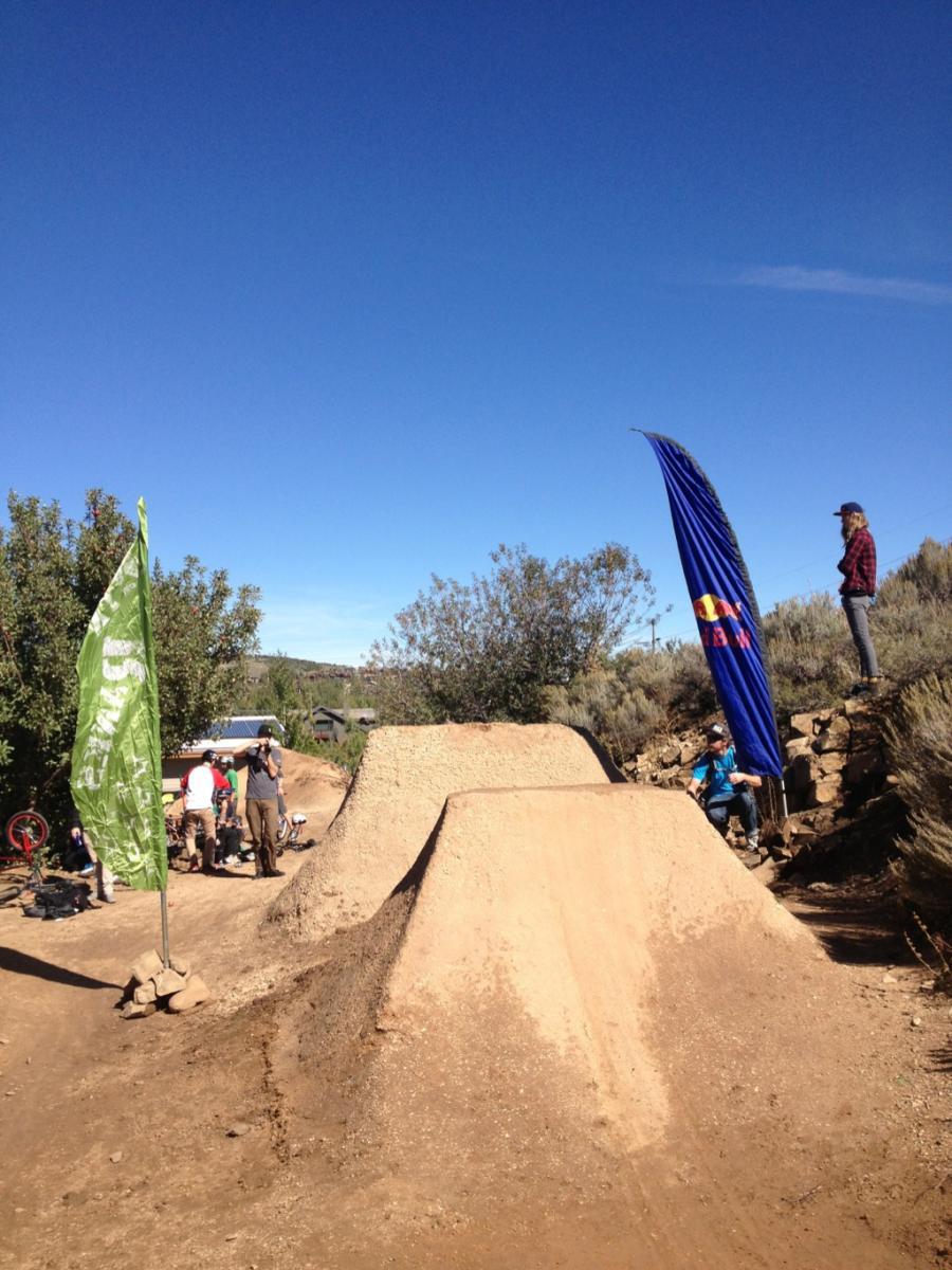 A dirt jump track surrounded by shrubs and trees, featuring two large dirt ramps and flags in the foreground. Several people are gathered nearby, some preparing to ride bikes, while a person stands atop one of the ramps. The sky is clear and blue, indicating a sunny day. PC Dirt Jumps mountain bike trail.