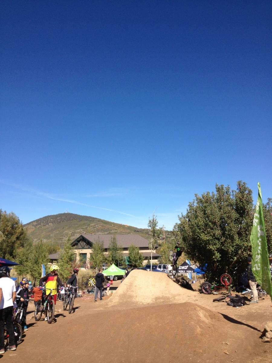 A bustling outdoor scene at a dirt bike park on a sunny day. Riders on bicycles are positioned around dirt jumps, with one rider performing a trick in the air. A crowd of spectators is gathered, and a building and trees are visible in the background under a clear blue sky. Flags and tents indicate an event or gathering taking place. PC Dirt Jumps mountain bike trail.
