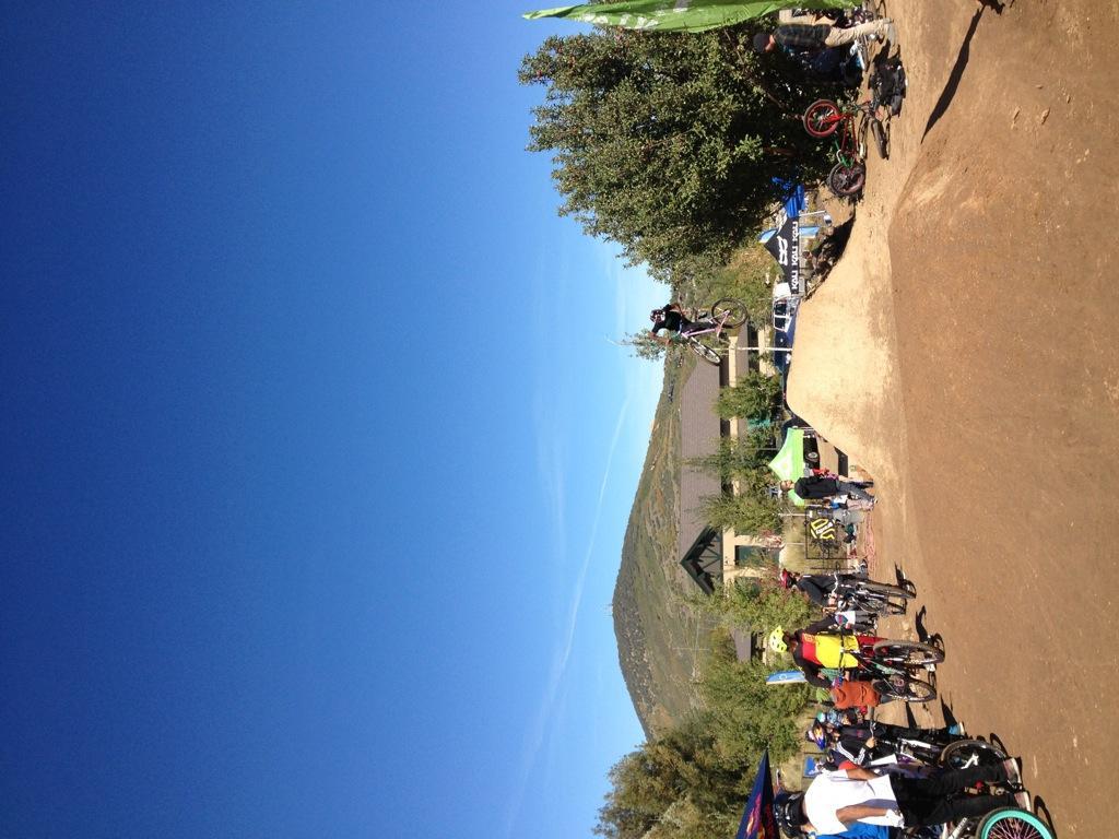 A dynamic scene at a bike park featuring a clear blue sky. A group of cyclists gathered around a dirt jump, while one rider is airborne, performing a trick above the jump. Spectators watch from the sidelines, and various tents and bikes are visible in the background. The landscape includes hills and trees, adding to the outdoor atmosphere. PC Dirt Jumps mountain bike trail.