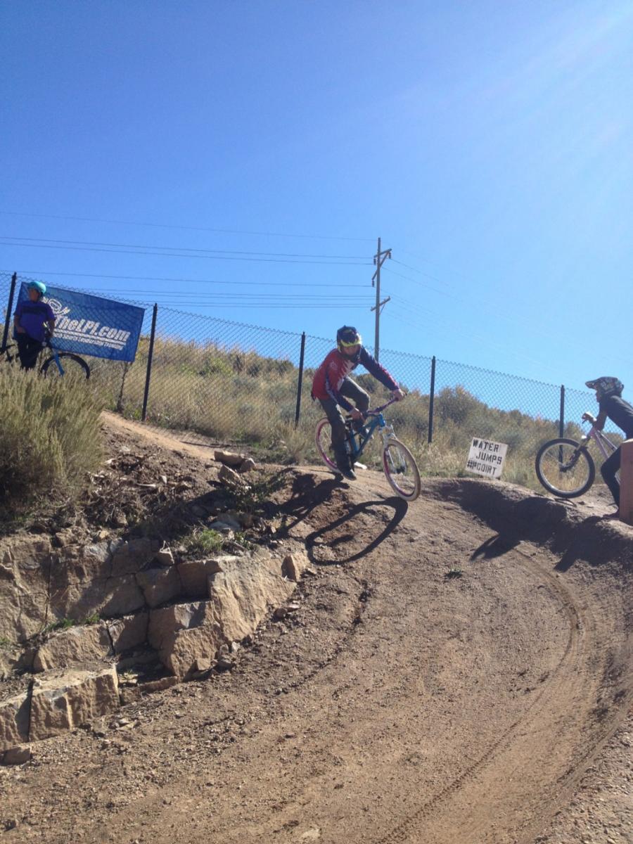 A mountain biker in a red jersey performs a jump on a dirt track, with another cyclist in a blue shirt nearby. The background features a blue sky and a fence with a sign that reads "WATER JUMPS HEADPOINT." The terrain is rugged with rocky landscaping and tire tracks visible on the path. PC Dirt Jumps mountain bike trail.