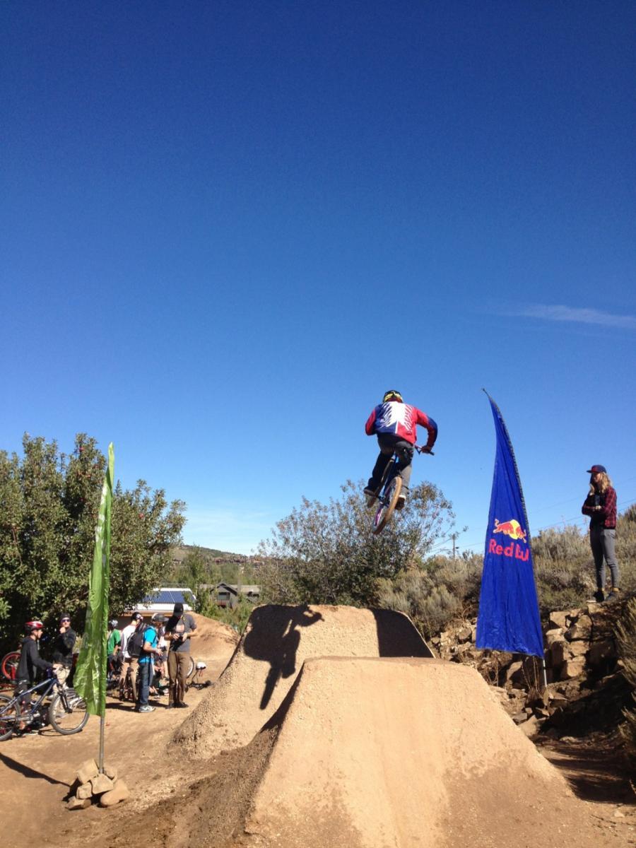 A mountain biker performing a jump over a dirt ramp at a biking event, with spectators watching nearby. Bright blue sky in the background and flags indicating event sponsorship are visible. PC Dirt Jumps mountain bike trail.