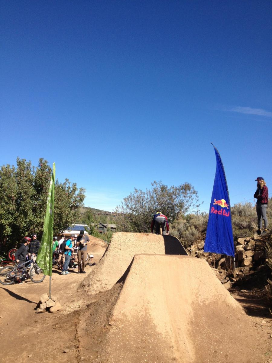 A dirt bike park scene featuring multiple riders on bikes, with one cyclist preparing to jump off a dirt ramp. Two flags are visible; one green and one blue with a Red Bull logo. Lush greenery surrounds the area, and the sky is clear and blue, indicating a sunny day. A group of people gathers nearby, watching the activity. PC Dirt Jumps mountain bike trail.
