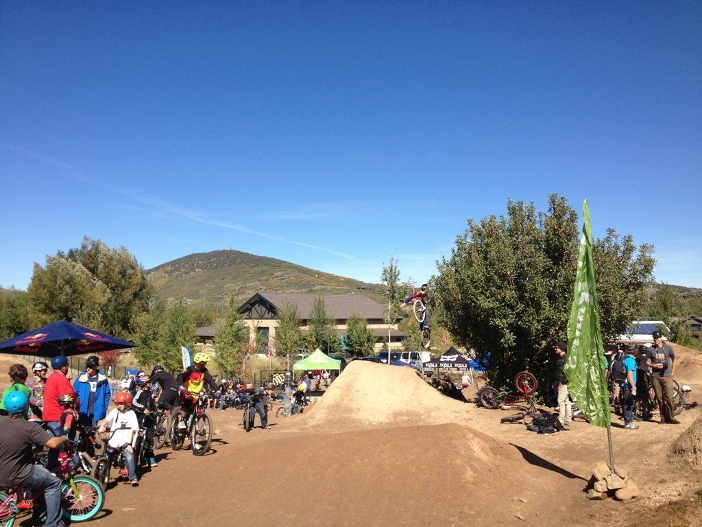 A lively outdoor scene at a bike park featuring a crowd of spectators watching a BMX rider performing a jump over a dirt ramp. The background includes green trees and buildings, with a clear blue sky overhead. Various bicycles can be seen, and flags and tents are set up around the area. PC Dirt Jumps mountain bike trail.