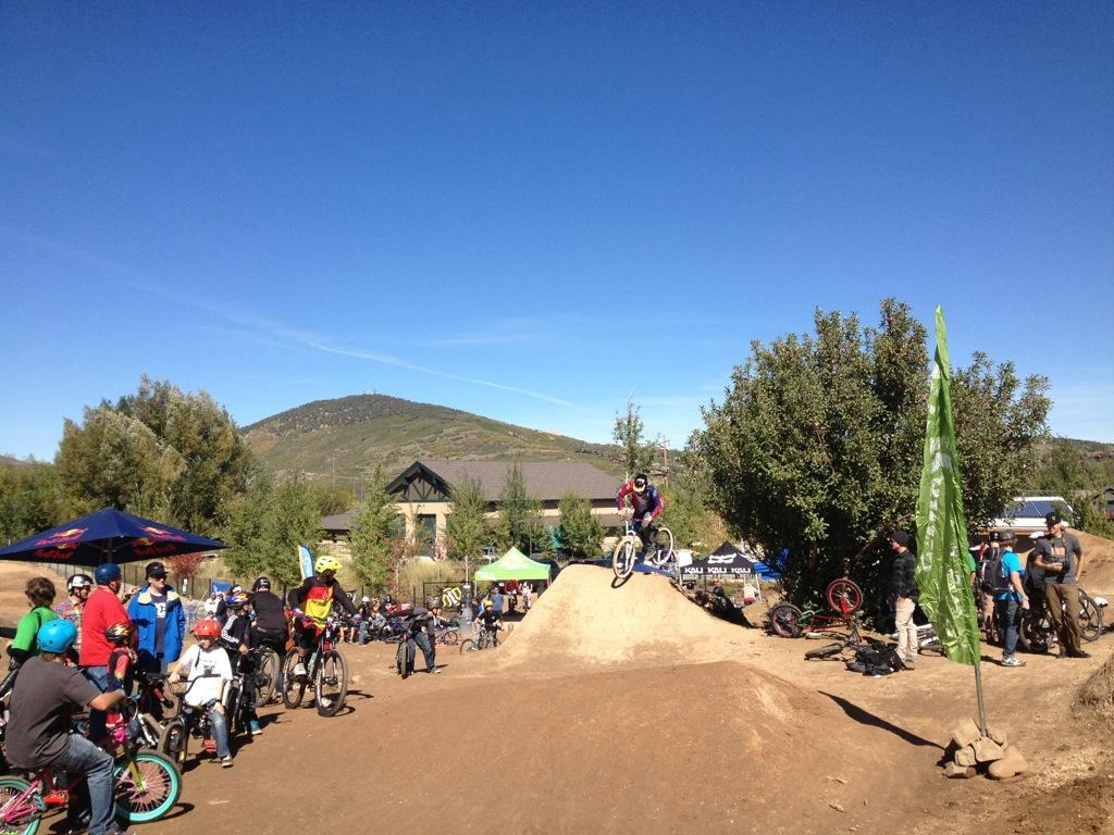 A sunny outdoor scene at a bike park featuring a dirt jump area. A cyclist in mid-air performs a jump over a dirt mound while a crowd of spectators and fellow riders watches nearby. Trees and flags are present, and a mountain can be seen in the background, highlighting a vibrant recreational atmosphere. PC Dirt Jumps mountain bike trail.