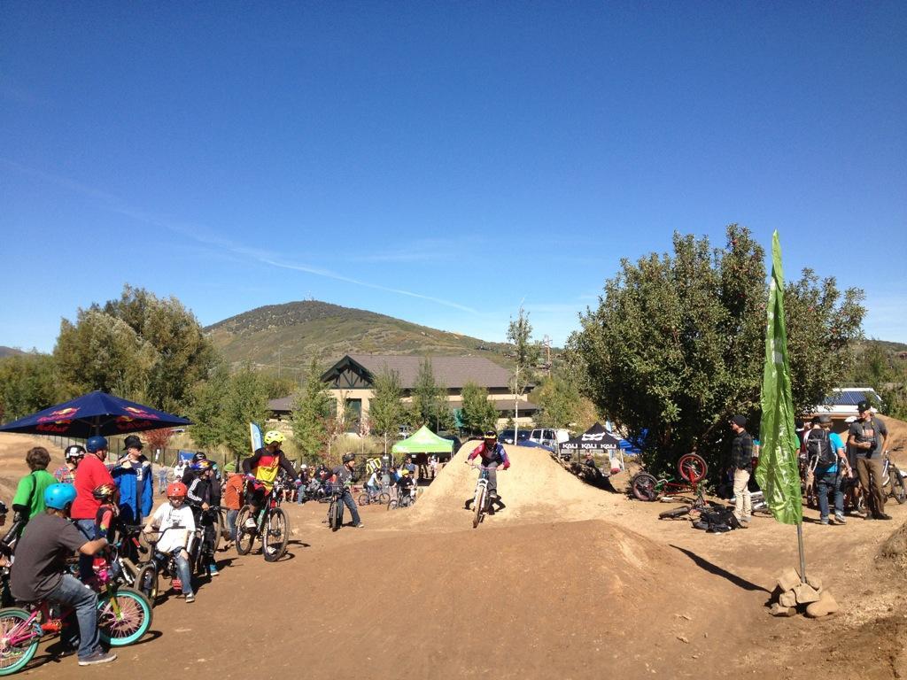 A vibrant outdoor scene at a bike park, showing a group of people gathered around a dirt jumping area. Several riders are taking turns performing jumps on their bicycles, while others watch from the sidelines. The background features a clear blue sky and rolling hills, with trees and a building visible nearby. Spectators include children and adults, all enjoying a sunny day of biking activities. PC Dirt Jumps mountain bike trail.