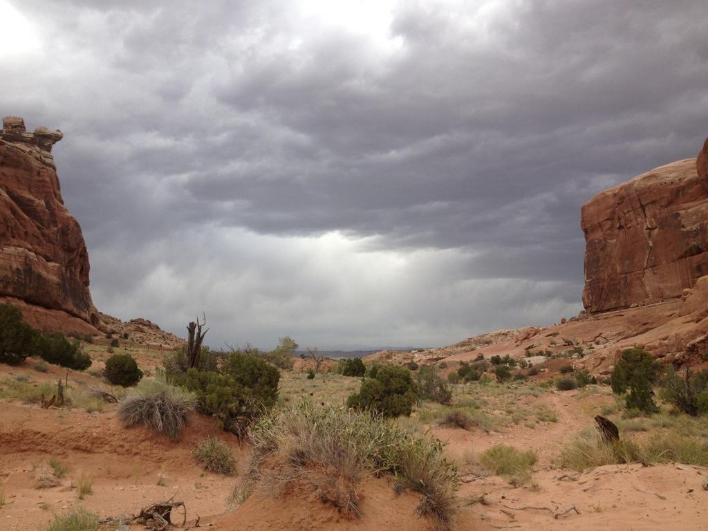 A rocky canyon with towering reddish-brown cliffs on either side, under a dramatic sky filled with dark clouds. The foreground features sparse vegetation and sandy terrain, leading into the canyon pathway. Monitor & Merrimac Singletrack mountain bike trail.