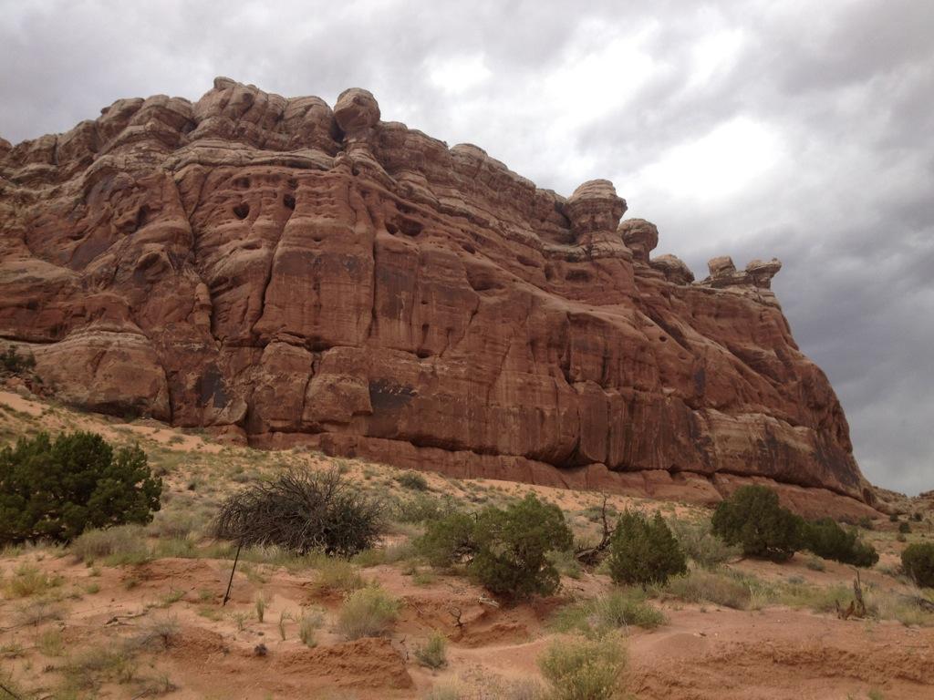 A dramatic rock formation stands tall against a cloudy sky, showcasing rugged layers of sandstone. The foreground features desert vegetation, including small bushes and sparse grass, set against a sandy ground. The scene captures a natural landscape characterized by its unique geological features. Monitor & Merrimac Singletrack mountain bike trail.