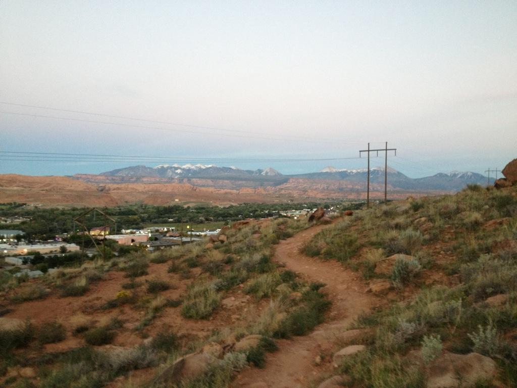 Scenic view of a winding dirt path leading through grassy terrain, with a valley and distant mountains visible under a twilight sky. Power lines run parallel to the path, while the foreground features a mix of shrubs and rocky outcrops. Pipe Dream mountain bike trail.