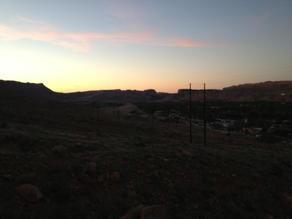 A landscape at dusk featuring rolling hills and distant mountains under a twilight sky with soft pastel colors. Silhouetted power poles stand on the hillside, overlooking a valley with scattered houses and vegetation. Pipe Dream mountain bike trail.