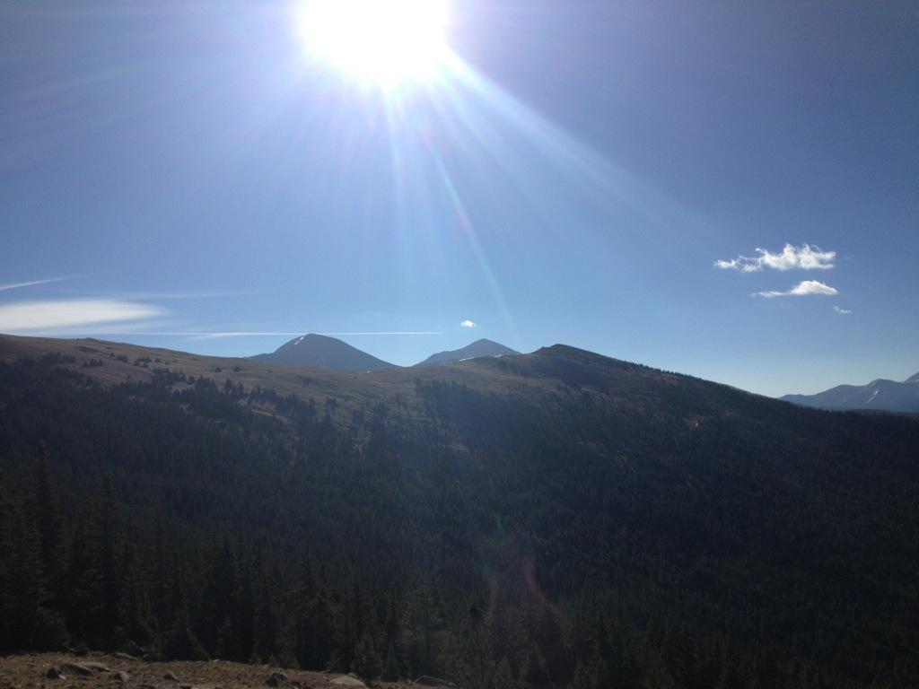 A panoramic view of a mountainous landscape under a clear blue sky, with the sun shining brightly in the top left corner. In the foreground, dense green forests cover the lower slopes, while the peaks of the mountains, some capped with snow, rise majestically in the background. A few wispy clouds are visible in the sky. Monarch Crest Trail mountain bike trail.