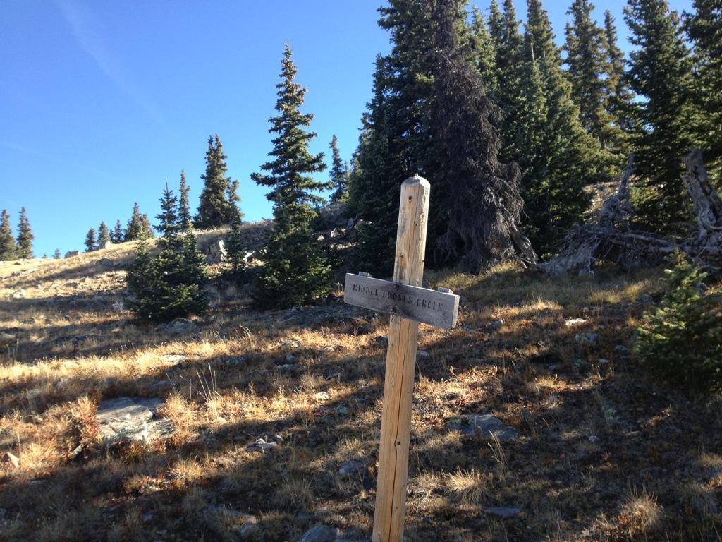 Wooden signpost marking the trail to "Kiddle Moosies Creek," surrounded by grassy terrain and evergreen trees under a clear blue sky. Monarch Crest Trail mountain bike trail.