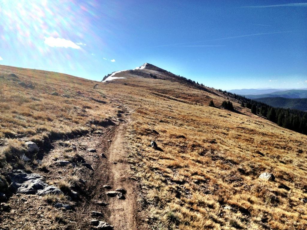 A wide view of a mountain trail leading up a grassy slope, with a clear blue sky above and scattered rocks along the path. In the background, the peak of the mountain is visible, partially covered with snow. The landscape features gentle hills and distant mountains under sunlight. Monarch Crest Trail mountain bike trail.