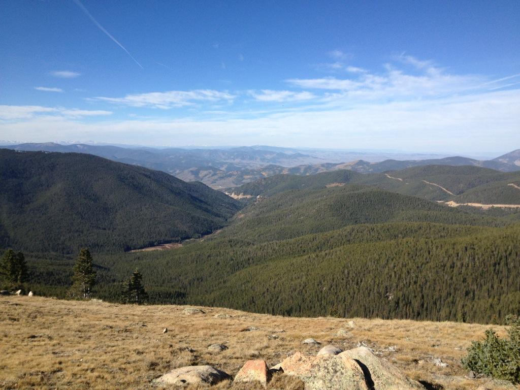 A panoramic view of rolling green mountains under a clear blue sky, with scattered clouds and a distant horizon. The foreground features rocky outcrops and patches of dry grass, while the landscape extends into layers of lush forested hills. Monarch Crest Trail mountain bike trail.