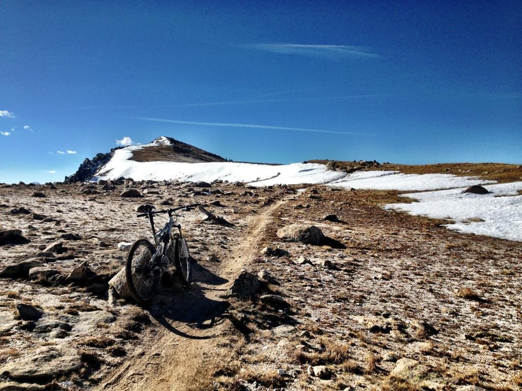 A mountain bike is parked on a rocky trail leading up a sunlit, snow-capped mountain peak under a clear blue sky. The landscape features a mix of rugged terrain and scattered snow patches. Monarch Crest Trail mountain bike trail.