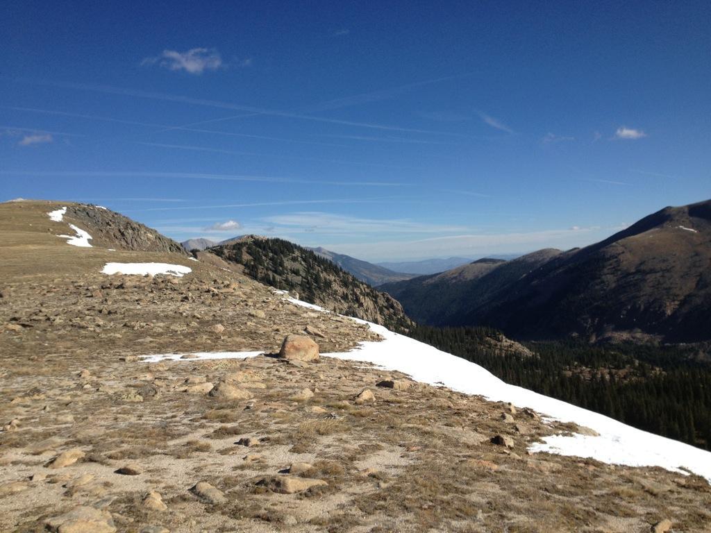 A panoramic view of mountainous terrain featuring rocky slopes and patches of snow under a clear blue sky. The landscape includes distant peaks and a valley with coniferous trees, showcasing the rugged beauty of the wilderness. Monarch Crest Trail mountain bike trail.