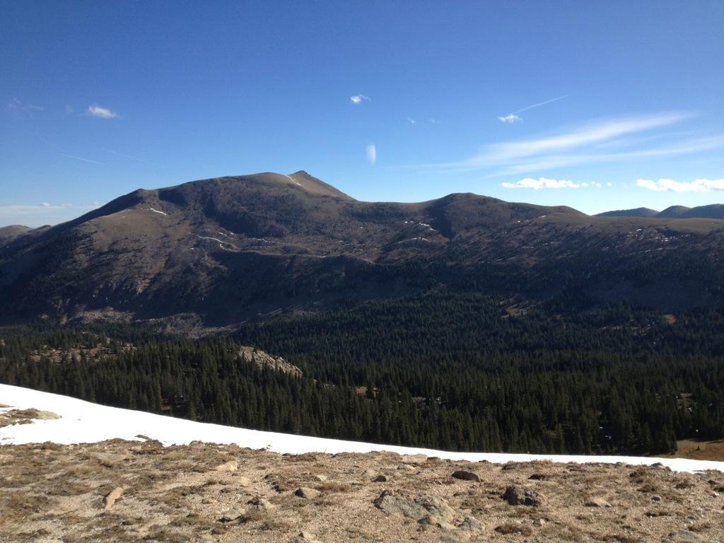 A panoramic view of a mountainous landscape under a clear blue sky, featuring rolling hills with sparse snow patches. The foreground includes rocky terrain and a small amount of snow, while the background showcases a dense forest of coniferous trees at the base of the mountains. The scene conveys a sense of tranquility and natural beauty. Monarch Crest Trail mountain bike trail.