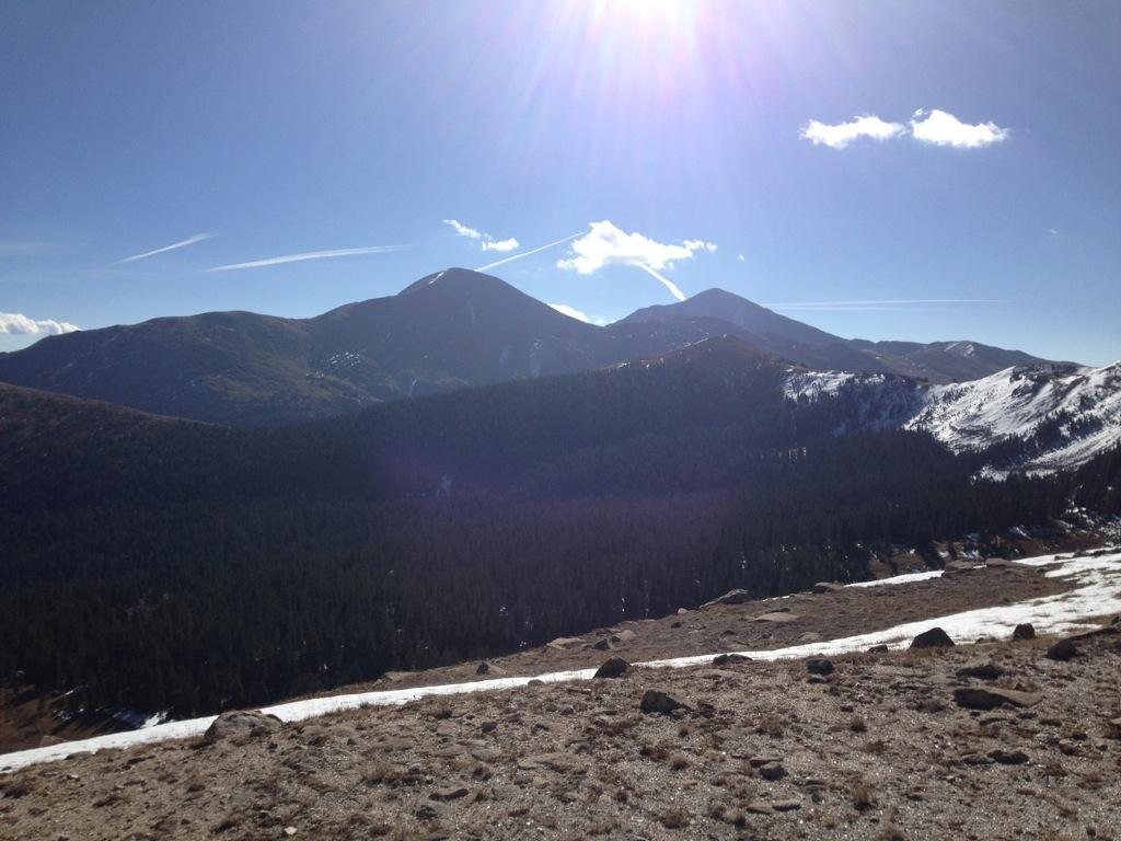 "Scenic view of a mountainous landscape under a clear blue sky, with sunlight shining over the peaks and a dense forest in the valley below. Snow-capped areas are visible on the mountains, highlighting the natural beauty of the terrain." Monarch Crest Trail mountain bike trail.