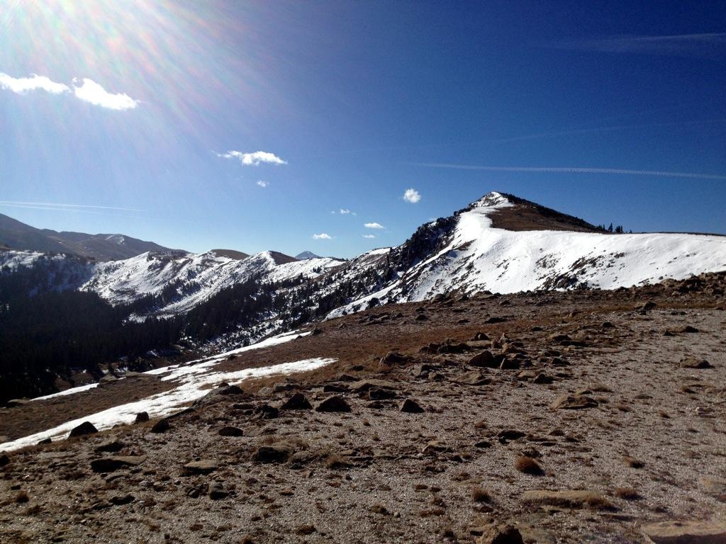A scenic mountain landscape featuring snow-capped peaks under a clear blue sky. The foreground shows rocky terrain and patches of snow, while the background displays rolling mountains with trees nestled in the valley. Sunlight creates a lens flare effect in the upper left corner. Monarch Crest Trail mountain bike trail.