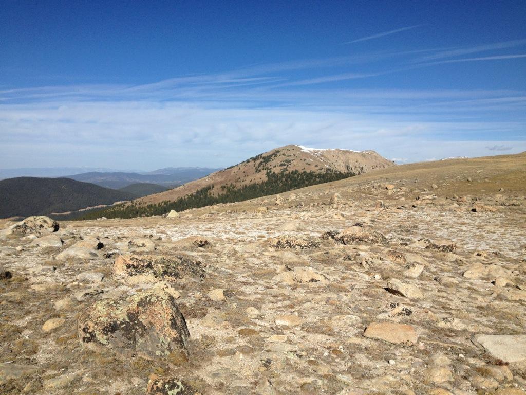 A wide-angle view of a rocky landscape on a mountain, featuring a gentle slope leading to a distant peak. The ground is covered with stones and sparse vegetation, with patches of snow visible on the mountain in the background. The sky is clear with light clouds, suggesting a bright day in a natural outdoor setting. Monarch Crest Trail mountain bike trail.