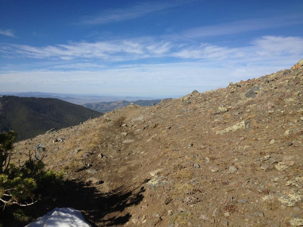A scenic view from a mountain ridge, featuring a rocky and sparse terrain under a clear blue sky with wispy clouds. Distant rolling hills and valleys are visible in the background, emphasizing the natural landscape. Monarch Crest Trail mountain bike trail.