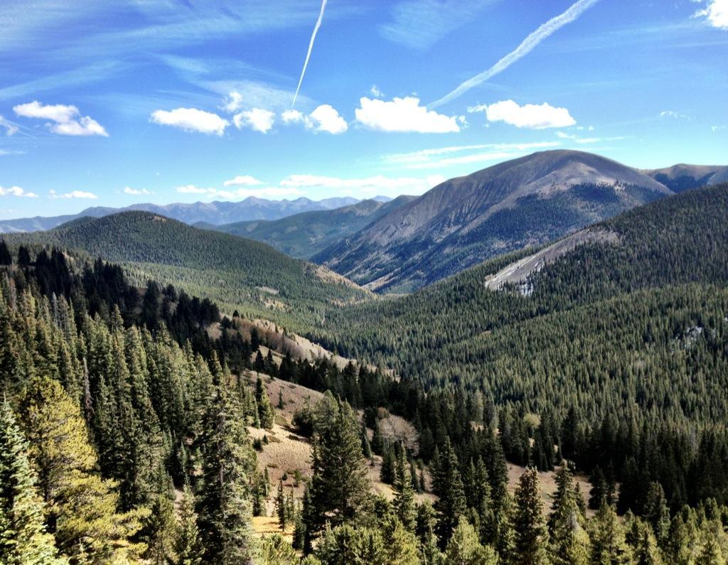 A panoramic view of a mountainous landscape featuring lush green forests, rolling hills, and distant mountain peaks under a blue sky with a few wispy clouds. A contrail is visible stretching across the sky. Monarch Crest Trail mountain bike trail.