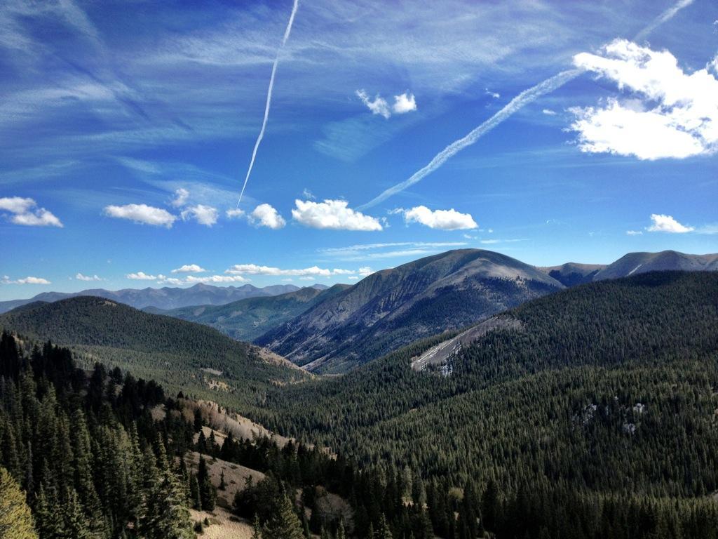 A wide view of a mountainous landscape under a blue sky with scattered white clouds. The scene features green forests covering the lower slopes, transitioning to rocky peaks in the background. Contrails from airplanes streak across the sky, adding to the serene, natural beauty of the area. Monarch Crest Trail mountain bike trail.