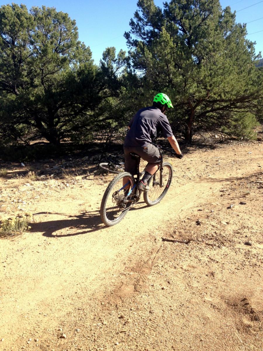 A person riding a mountain bike on a dirt trail surrounded by trees, wearing a green helmet and a gray shirt. Sunlight shines on the path, highlighting the textured ground. Little Rainbow mountain bike trail.
