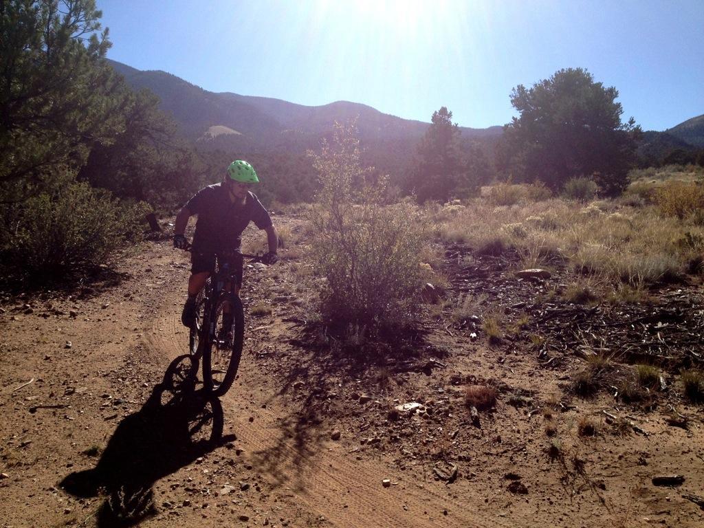 A person riding a mountain bike along a dirt trail in a sunlit outdoor setting, surrounded by shrubs and trees, with mountains in the background. The cyclist is wearing a green helmet and a dark outfit, and the terrain is rocky and sandy. Little Rainbow mountain bike trail.