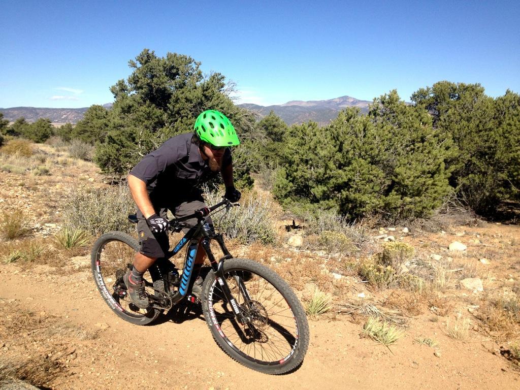 A mountain biker in a green helmet navigates a dirt trail surrounded by shrubs and trees, with distant mountains visible in the background under a clear blue sky. Little Rainbow mountain bike trail.
