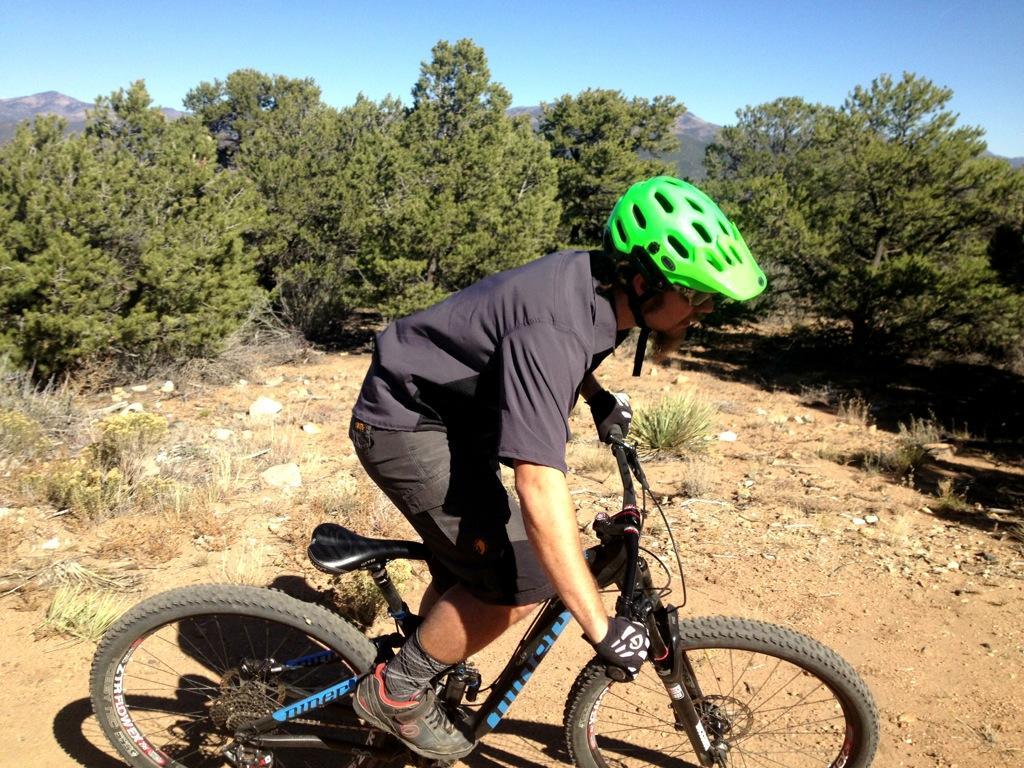A cyclist wearing a bright green helmet rides a mountain bike along a dirt trail surrounded by pine trees and rocky terrain under a clear blue sky. Little Rainbow mountain bike trail.