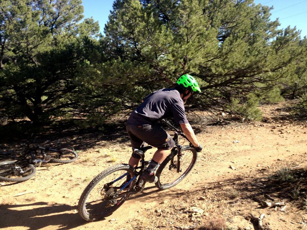 A person riding a mountain bike on a dirt trail surrounded by trees. The rider is wearing a bright green helmet and casual biking attire, leaning slightly forward as they navigate the path. In the background, another bike is visible resting on the ground among the foliage. Little Rainbow mountain bike trail.