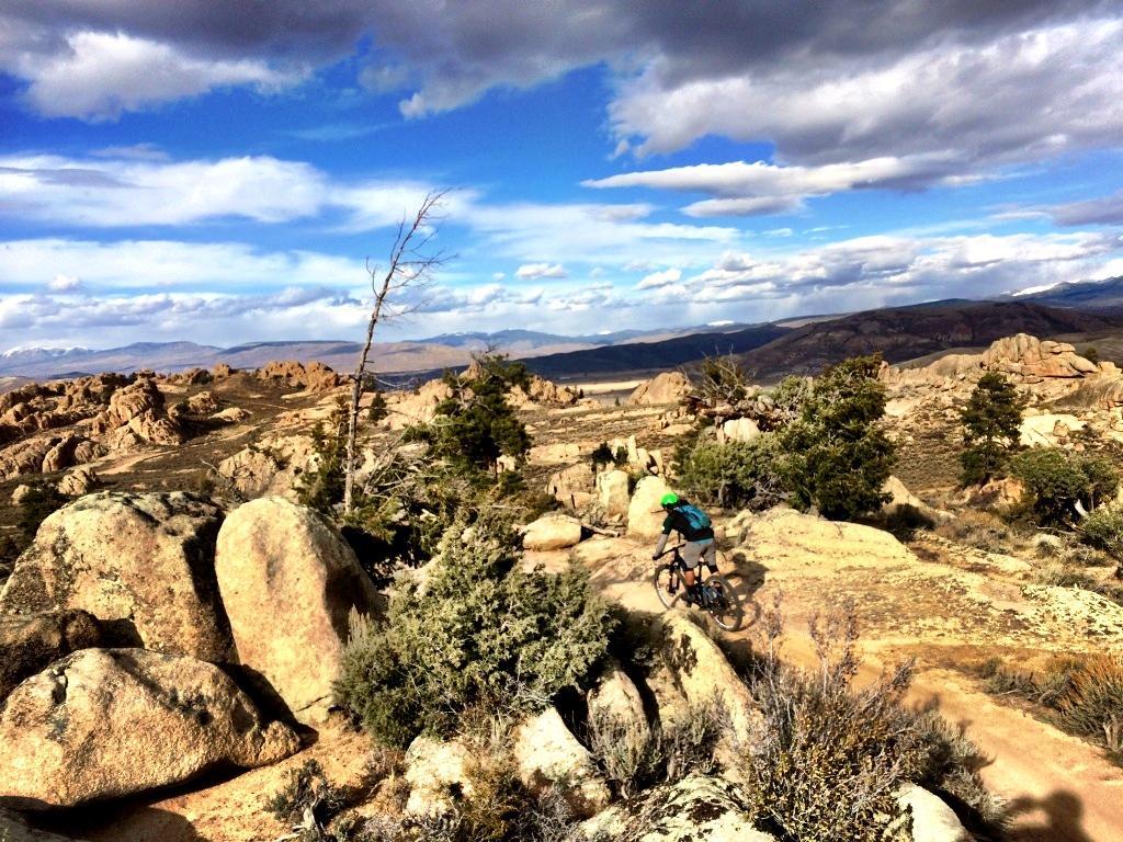 A mountain biker navigating a rocky trail in a scenic outdoor landscape, featuring rugged boulders and sparse vegetation under a bright blue sky with scattered clouds. Background includes distant mountains and an expansive view of the terrain. Hartman Rocks mountain bike trail.