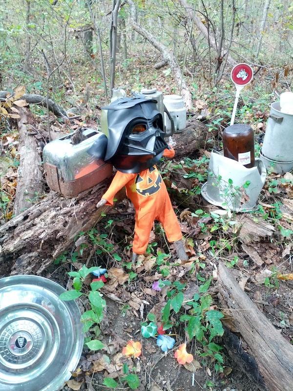 A small doll dressed in an orange pumpkin-themed outfit and wearing a black mask stands among fallen branches in a wooded area. Surrounding the doll are various vintage items, including a shiny metal object, a glass jar, and a sign. Colorful plastic flowers are scattered on the ground nearby, adding a whimsical touch to the scene. Rosaryville State Park mountain bike trail.