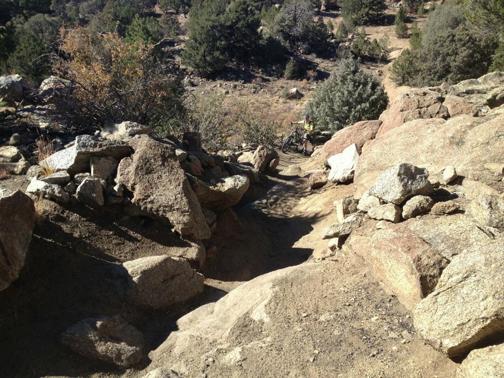 A rocky mountain biking trail featuring large boulders and loose dirt, surrounded by trees and shrubs. A cyclist is visible in the background, standing near their bike. Midland Hills Trails mountain bike trail.