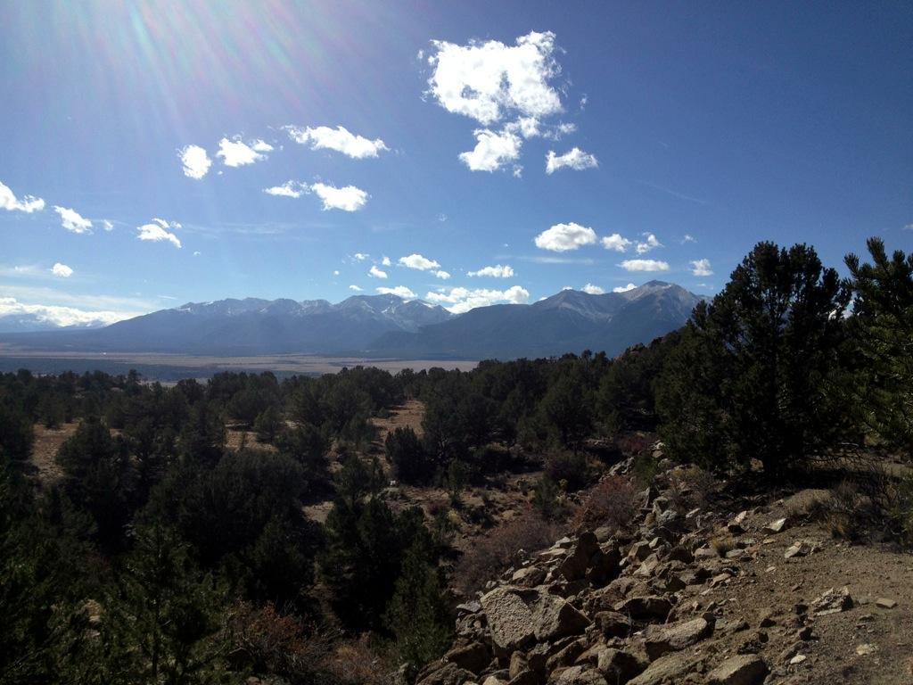 Scenic view of a mountainous landscape featuring snow-capped peaks under a blue sky with fluffy white clouds. In the foreground, a mix of evergreen trees and rocky terrain can be seen, leading to a valley below. Midland Hills Trails mountain bike trail.