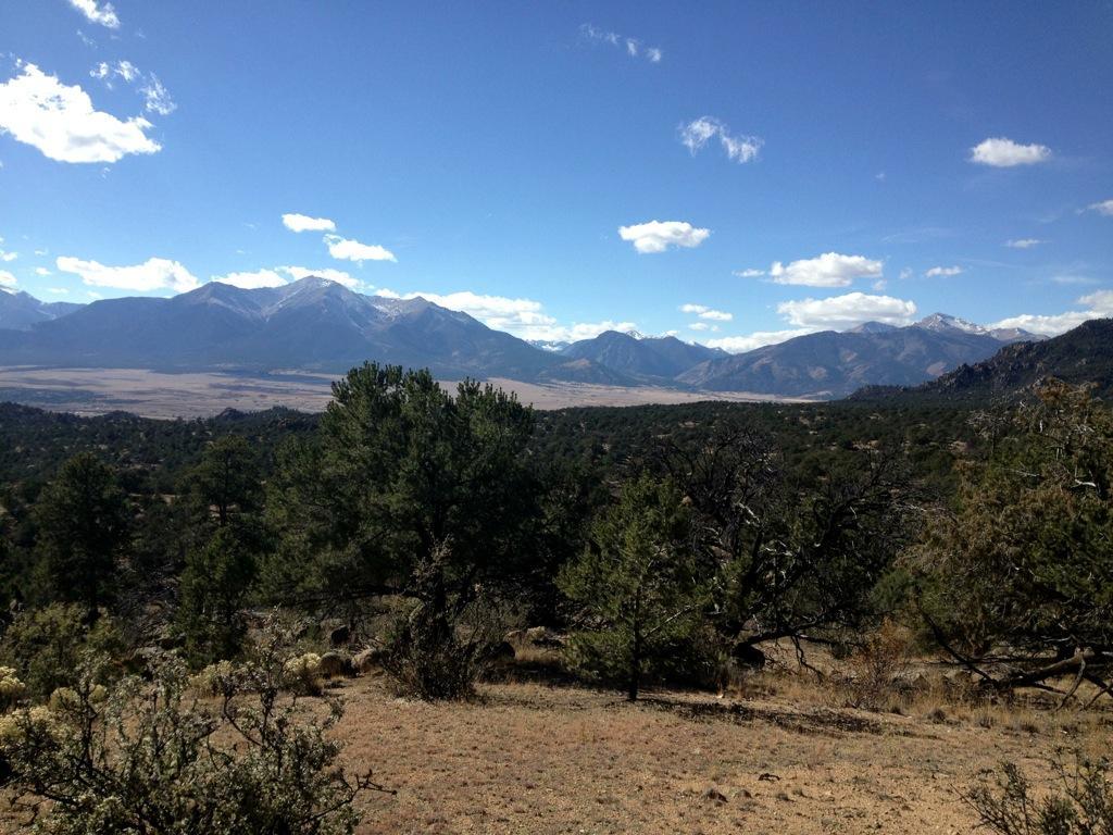 A panoramic view of a mountainous landscape featuring snow-capped peaks under a bright blue sky with scattered clouds. In the foreground, green trees and shrubs are visible, leading to a valley that stretches out towards the mountains in the distance. Midland Hills Trails mountain bike trail.