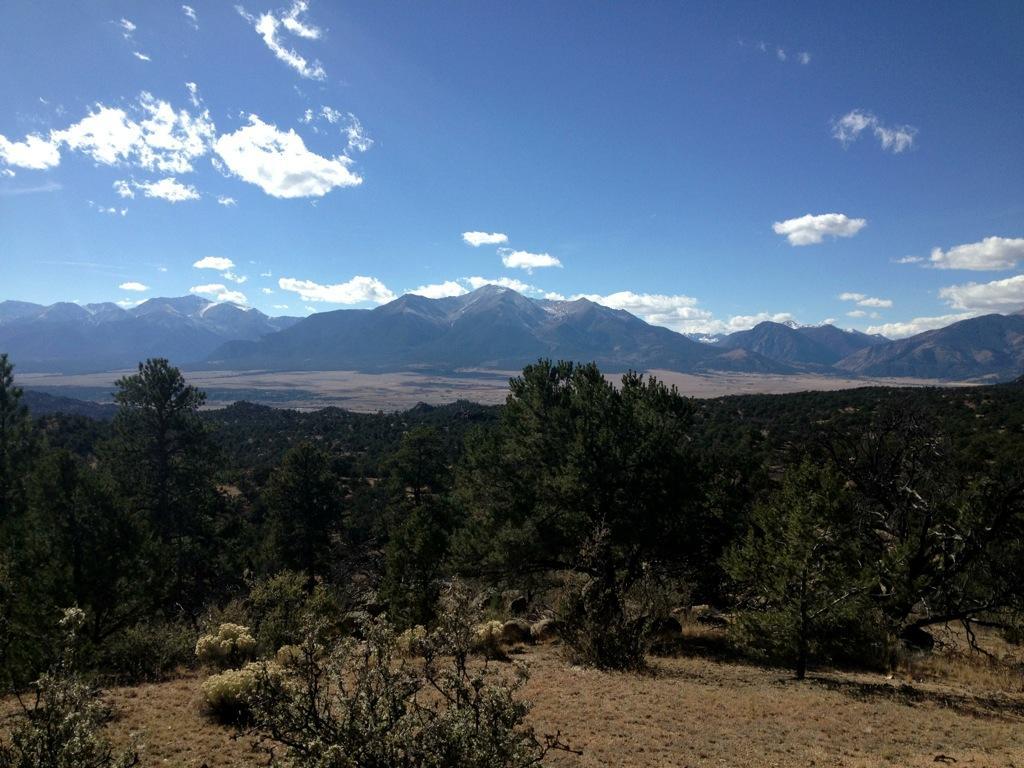 A panoramic view of a mountainous landscape under a blue sky, featuring distant snow-capped peaks and patches of clouds. In the foreground, there are scattered trees and shrubs, with a valley stretching out below, showcasing a mix of green vegetation and dry terrain. Midland Hills Trails mountain bike trail.