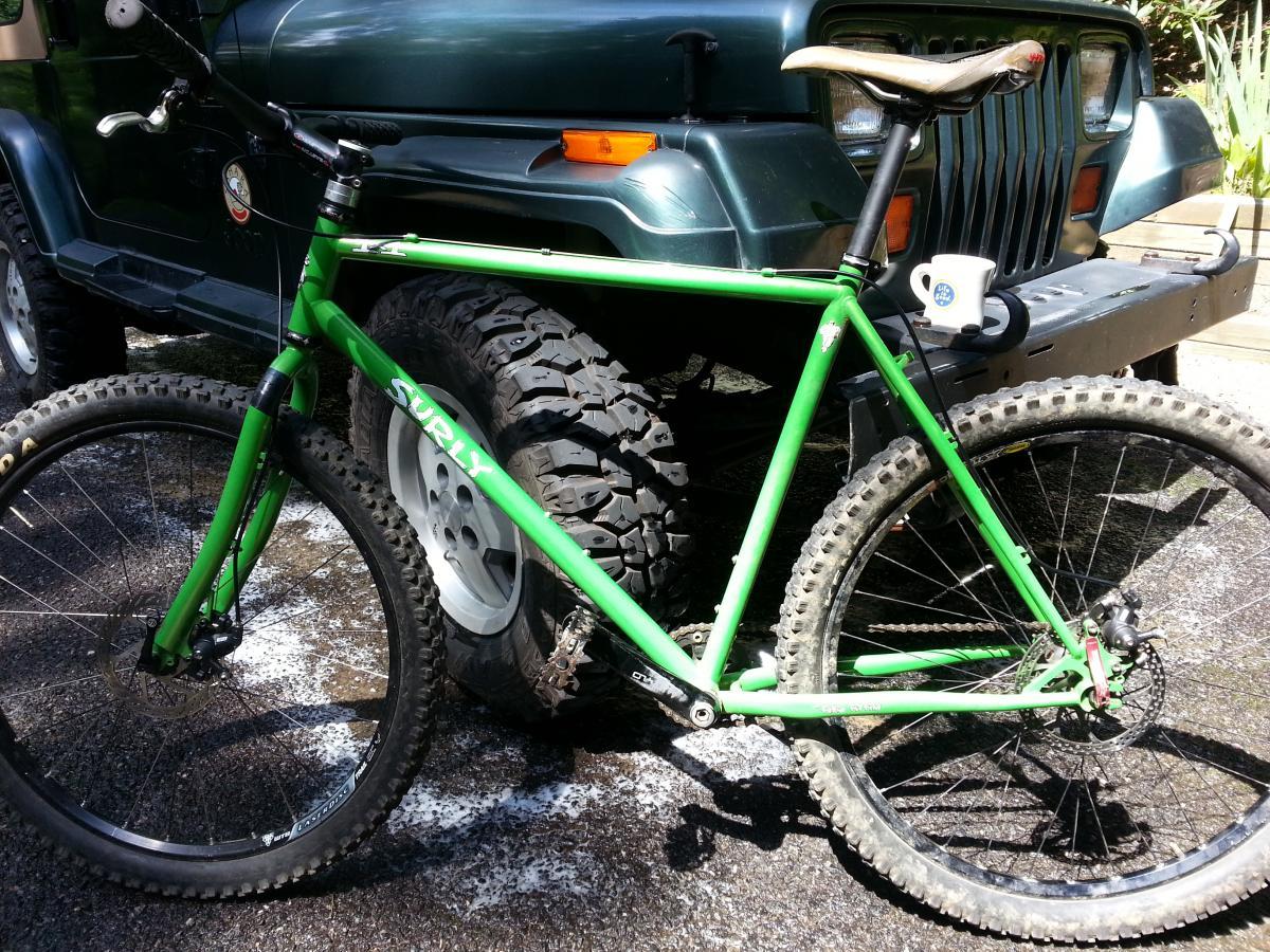 Specialized Stumpjumper FSR Comp 29: A green Surly mountain bike parked next to a dark-colored Jeep, showcasing the bike's distinctive frame and rugged tires on a paved surface. A small coffee cup is resting on the Jeep's hood in the background.