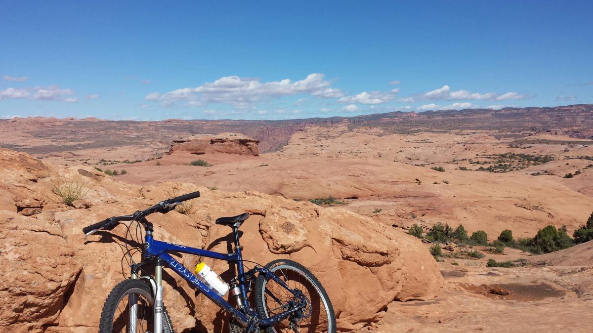 Trek Fuel EX 7: A mountain bike resting on a rocky outcrop overlooking a vast desert landscape with rolling hills and a blue sky dotted with fluffy white clouds.