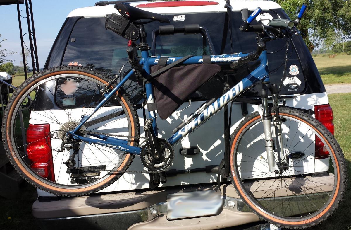 Specialized Rockhopper: A blue Specialized mountain bike mounted on the back of a white SUV. The bike features rugged tires and a small bag attached to the frame. In the background, a grassy area and trees are visible under a clear sky.