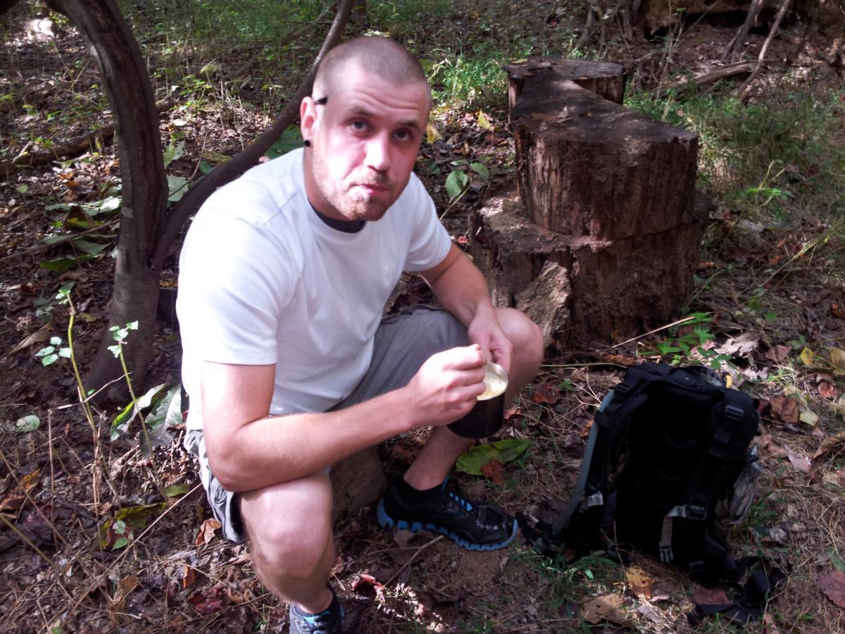 A person sitting on a stump in a forested area, holding a small container while eating. They are wearing a white t-shirt and shorts, with a backpack next to them. The ground is covered in fallen leaves and the background features trees and greenery. Turkey Creek mountain bike trail.