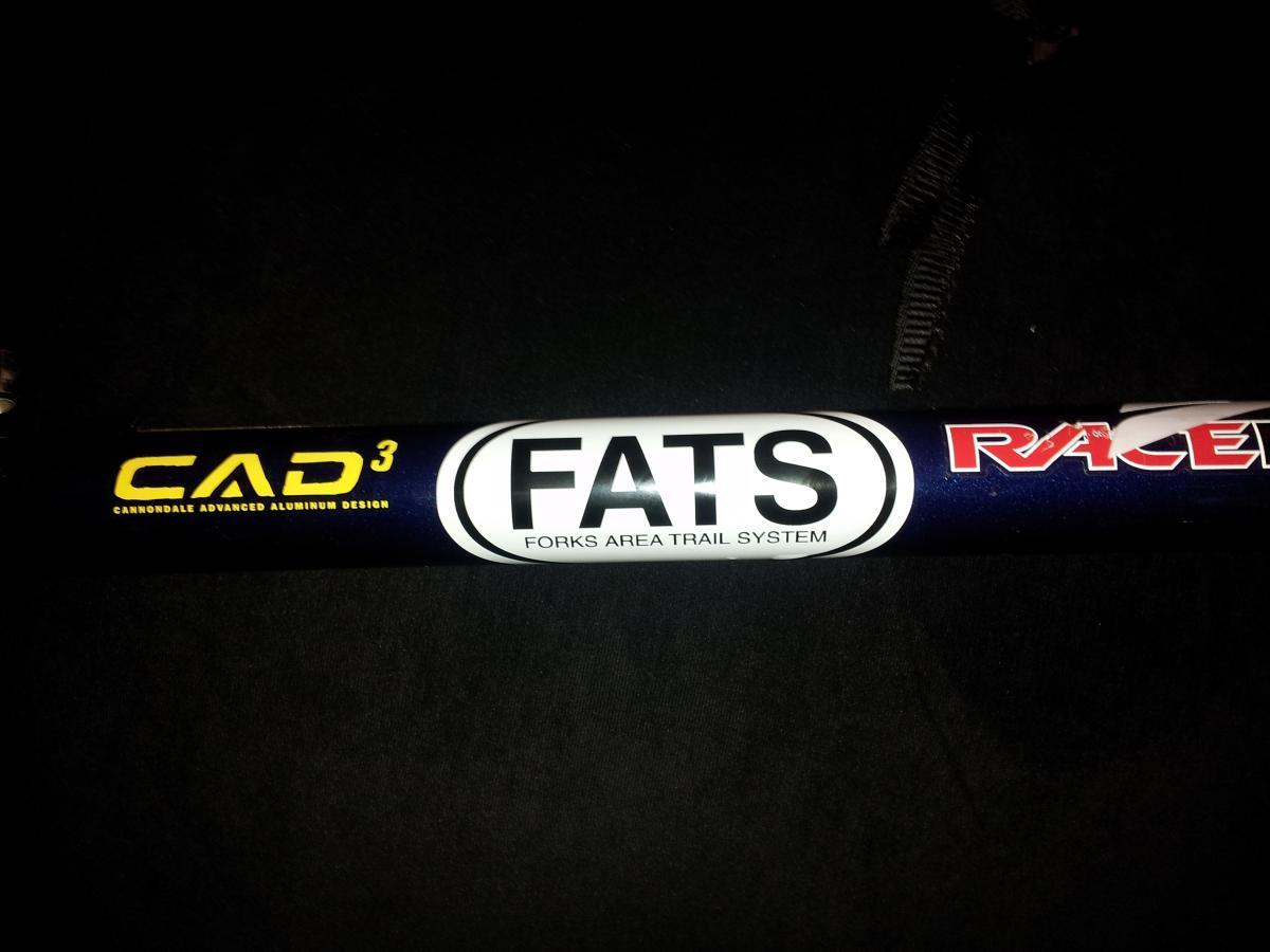 Image of a bicycle frame tube displaying decals that read "CAD³" in yellow, indicating Cannondale Advanced Aluminum Design, and "FATS" in bold black letters, representing the Forks Area Trail System, alongside the word "Racer" in red text. The background is dark, highlighting the details on the frame. Forks Area Trail System (FATS) mountain bike trail.
