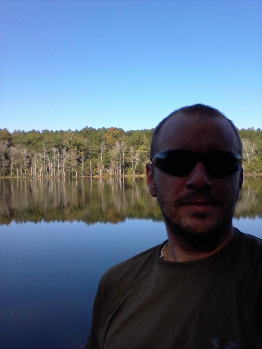 A man wearing sunglasses stands in front of a calm lake, surrounded by trees reflecting in the water. The sky is clear and blue, indicating a sunny day. Boyd Pond mountain bike trail.