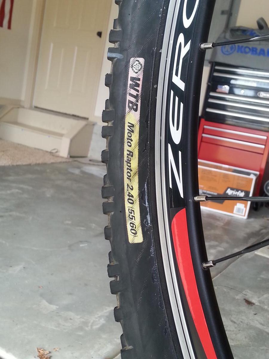 WTB Moto Raptor: Close-up view of a mountain bike tire featuring the WTB Moto Raptor model label, displaying tire specifications including a width of 2.40 and diameter of 26 inches. The background is a garage setting with tools and equipment visible.