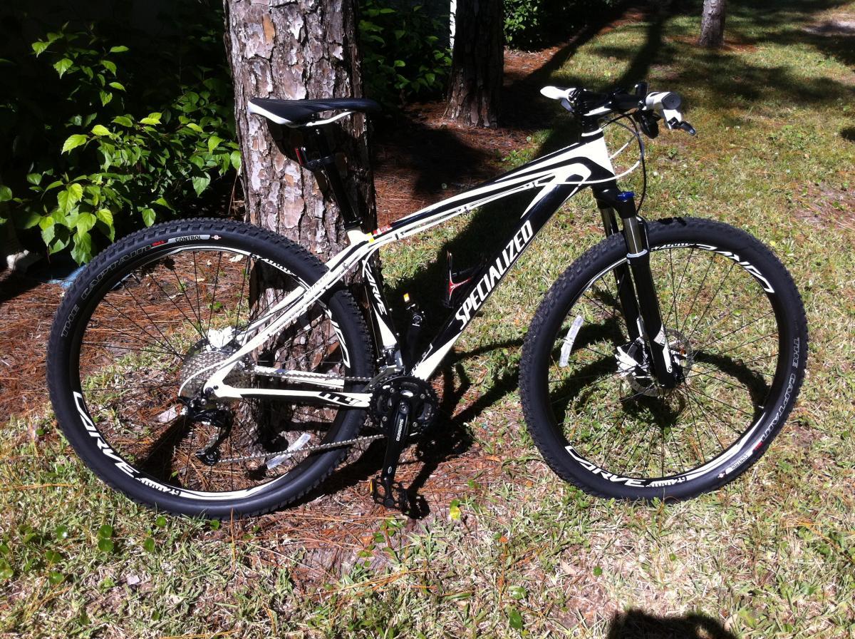 Specialized Carve Expert 29er: A white and black Specialized mountain bike leaning against a tree, set in a grassy area with foliage in the background. The bike features large tires and a lightweight frame, designed for off-road cycling.