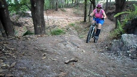 A person riding a mountain bike on a dirt trail surrounded by trees, with patches of leaves and rocks in the background. The rider is wearing a pink helmet, a pink top, and blue shorts, navigating a slope in a wooded area. Balm Boyette Scrub Preserve mountain bike trail.